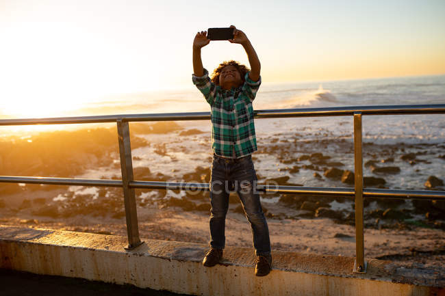 Front view of a smiling pre-teen boy holding a smartphone over his head and taking a selfie leaning on a balustrade at a sunset by the sea — Stock Photo
