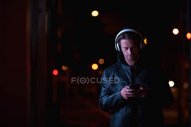 Вид спереди на молодого кавказца, стоящего вечером на улице, слушающего музыку в наушниках, держащего смартфон — стоковое фото
