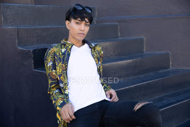 Porträt eines modischen jungen Transgender-Erwachsenen mit gemischter Rasse auf der Straße, der auf Stufen an einer grauen Wand sitzt — Stockfoto