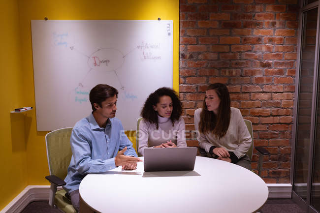 Frontansicht einer jungen Frau mit gemischter Rasse und einer jungen kaukasischen Frau und einem Mann, die an einem Tisch im Gespräch um einen Laptop sitzen und gemeinsam im Büro eines kreativen Unternehmens arbeiten — Stockfoto
