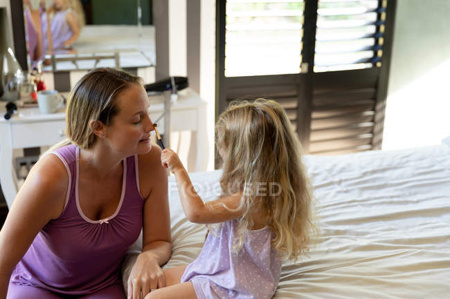 Vista lateral de una joven embarazada caucásica maquillada por su hija en su dormitorio - foto de stock