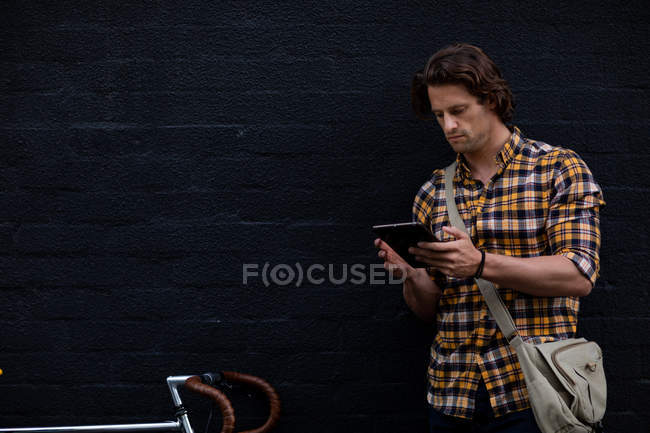 Vista frontal de um jovem caucasiano de pé na rua segurando um computador tablet com uma bicicleta ao lado dele durante sua viagem à noite para casa — Fotografia de Stock