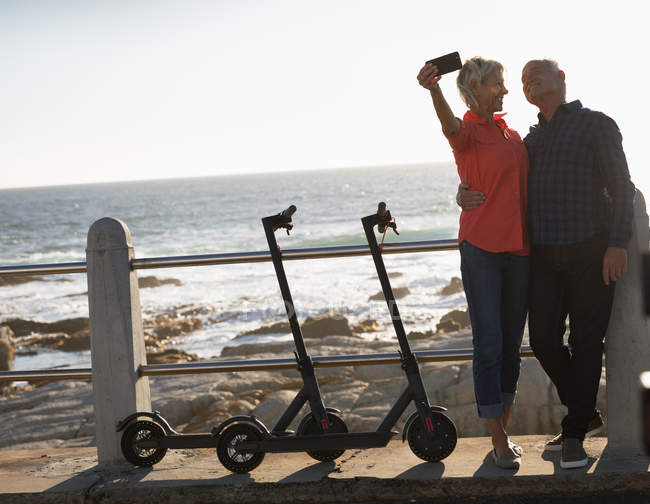 Front view of a mature Caucasian man and woman taking a selfie next to e scooters by the sea at sunset — Stock Photo