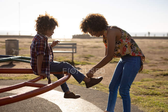 Vue latérale d'une jeune femme métissée et de son fils pré-adolescent jouissant du temps passé ensemble dans une aire de jeux au bord de la mer, assis sur un carrousel par une journée ensoleillée — Photo de stock