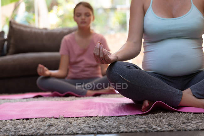 На передньому плані - молода кавказька вагітна жінка, що займається йогою зі своєю донькою у вітальні. — стокове фото