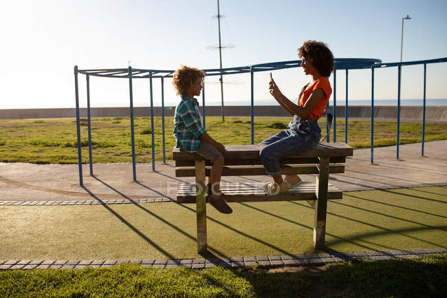 Вид сбоку на молодую расовую женщину и ее сына-подростка, играющих вместе на детской площадке у моря, сидящих на скамейке и фотографирующих в солнечный день на заднем плане — стоковое фото