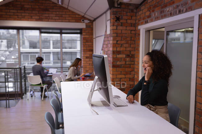Vista laterale da vicino di una giovane donna di razza mista seduta a una scrivania di fronte a un computer che parla con un auricolare nell'ufficio di un'azienda creativa, con colleghi seduti a lavorare sullo sfondo — Foto stock
