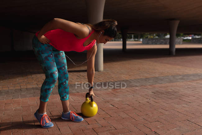 Side view of a young Caucasian woman wearing sports clothes bending forward with one hand on her hip and lifting a kettlebell weight with the other hand during a workout on a sunny day in a park — Stock Photo