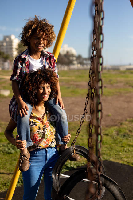 Front view close up of a young mixed race woman and her pre-teen son enjoying time together playing at a playground, the woman carries her son piggyback on a sunny day with swings in the foreground — Stock Photo