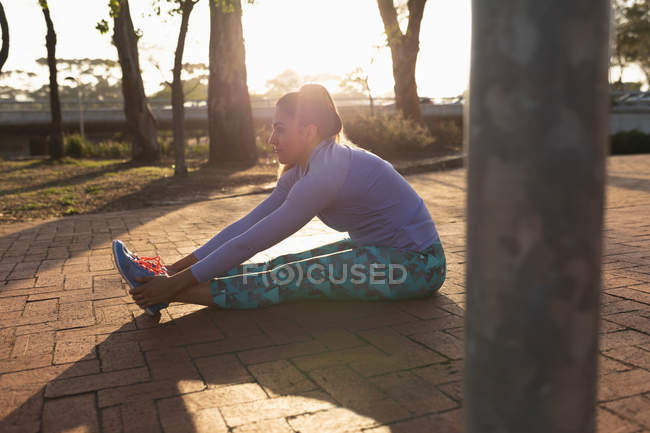 Side view of a young Caucasian woman wearing sports clothes sitting down and holding her ankles with legs stretched out in front of her while working out in a park — Stock Photo