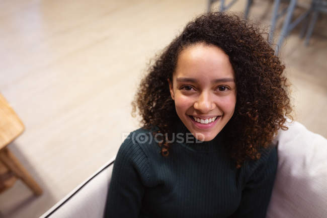 Ritratto da vicino di una giovane donna di razza mista che guarda verso la macchina fotografica e sorride, seduta su un divano nell'area salotto di un ufficio creativo — Foto stock