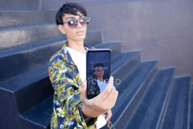 Vista frontal de um jovem elegante mestiço transexual adulto na rua, mostrando a tela do smartphone, tirando uma selfie — Fotografia de Stock