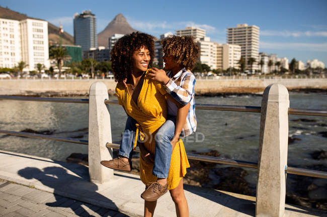 Frontansicht einer jungen Mischlingshündin und ihres vorpubertären Sohnes, die die gemeinsame Zeit am Meer genießen, trägt die Frau ihren Sohn huckepack an einem sonnigen Tag — Stockfoto