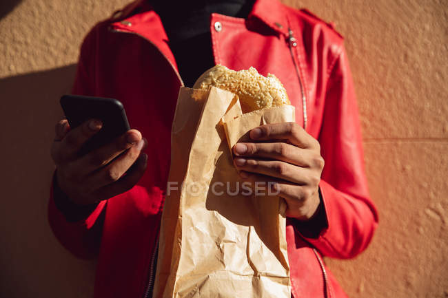 Vista frontal sección media de un hombre de moda en la calle, usando un teléfono inteligente y comiendo un sándwich - foto de stock