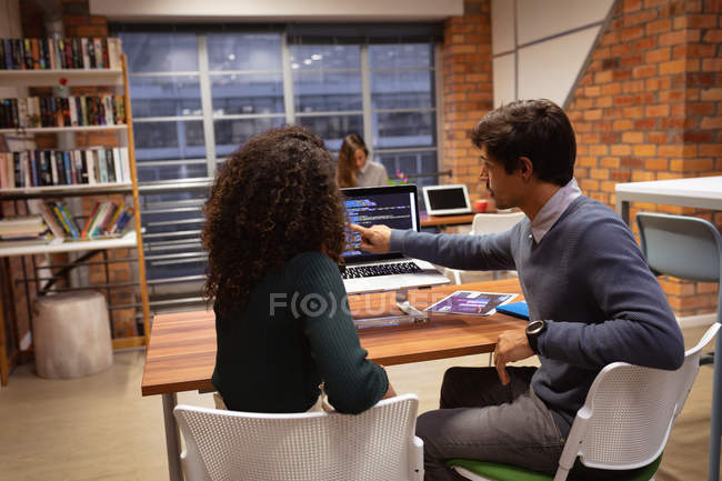 Зворотний вид молодого кавказького чоловіка і молодої жінки змішаної раси, яка сидить за столом за допомогою ноутбука і розмовляє в офісі творчого бізнесу, з колегою, що працює за столом на задньому плані. — стокове фото