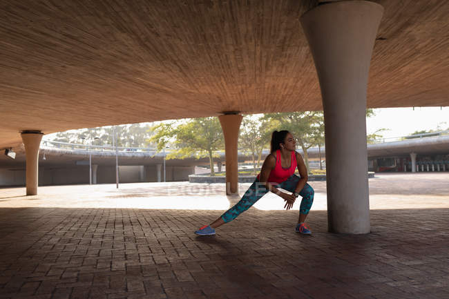 Vista frontale di una giovane donna caucasica che indossa vestiti sportivi accovacciati e si estende sotto un ponte durante un allenamento in un parco — Foto stock
