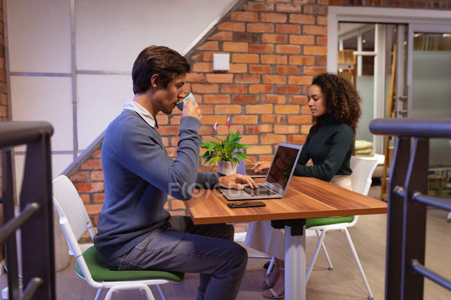 Side view close up of a young Caucasian man and a young mixed race woman sitting at a desk using laptop computers in the office of a creative business, the man is drinking a cup of coffee while he works — Stock Photo