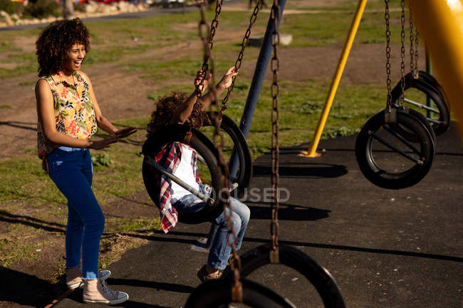 Vue latérale d'une jeune femme métissée et de son fils pré-adolescent jouissant du temps passé ensemble sur une aire de jeux, le garçon se balançant par une journée ensoleillée — Photo de stock