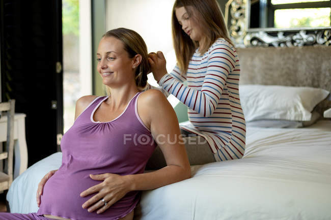 Vista laterale da vicino di una ragazza caucasica che spazzola i capelli della madre incinta nella sua camera da letto — Foto stock
