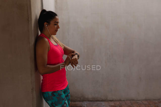 Vista lateral de una joven mujer caucásica con ropa deportiva apoyada en una pared revisando su reloj inteligente mientras hace ejercicio - foto de stock