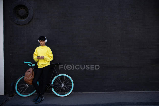 Vista frontal de un joven transexual de raza mixta de moda en la calle, mensajes de texto en el teléfono inteligente de pie junto a una bicicleta con auriculares en contra de una pared gris - foto de stock