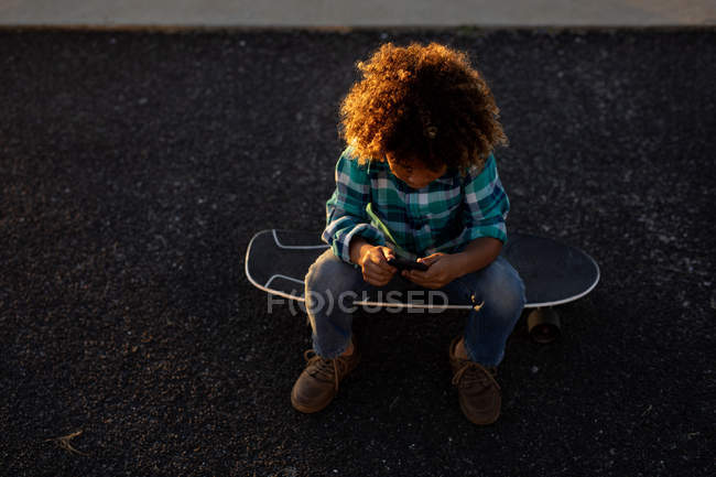 Hochwinkelaufnahme eines Jungen im Teenageralter, der mit einem Smartphone auf einem Skateboard sitzt — Stockfoto