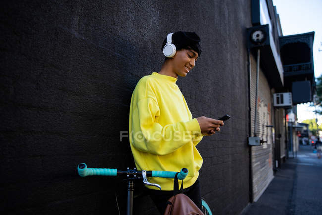 Vista lateral de un joven transexual de raza mixta de moda en la calle, mensajes de texto en el teléfono inteligente de pie junto a una bicicleta con auriculares - foto de stock