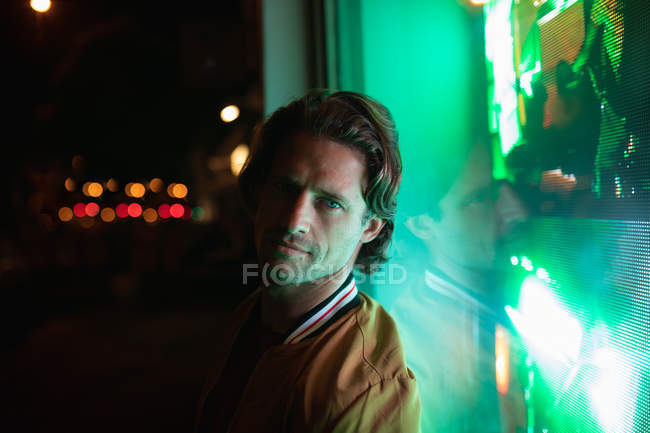 Portrait of a young Caucasian man looking to camera in the evening with green neon light from a shop window behind him — Stock Photo