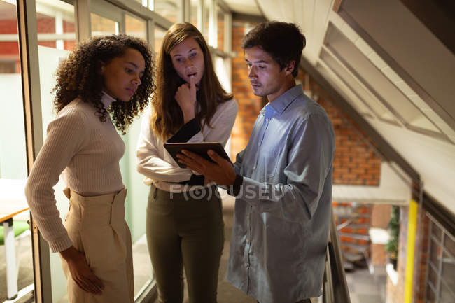 Seitenansicht einer jungen Frau mit gemischter Rasse und einer jungen kaukasischen Frau und einem Mann, die im Büro eines kreativen Unternehmens stehen und auf einen Tablet-Computer blicken, den der Mann zusammenhält — Stockfoto
