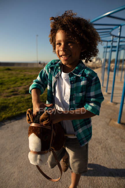 Vista frontal de cerca de un niño preadolescente de raza mixta jugando en un patio de recreo, de pie con un caballo hobby en un día soleado - foto de stock