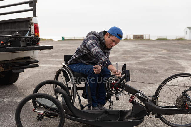 Вид спереди молодого кавказца в инвалидной коляске, собирающего лежачий велосипед, фиксирующего цепь на стоянке у моря — стоковое фото