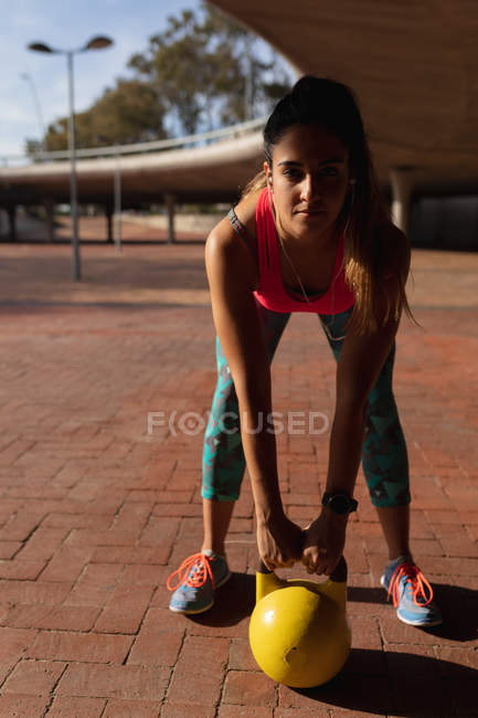 Front view close up of a young Caucasian woman wearing sports clothes leaning forward holding a kettlebell weight during a workout on a sunny day in a park — Stock Photo