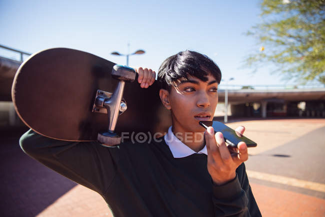Visão frontal seção média de um jovem elegante mestiço transexual adulto na rua, falando no smartphone e segurando um skate — Fotografia de Stock