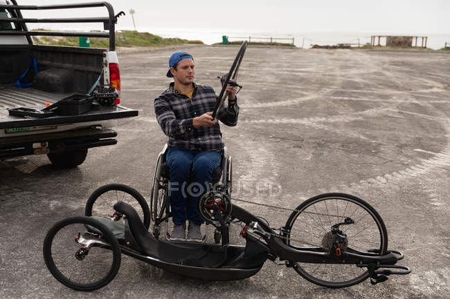 Vista frontal de um jovem caucasiano em uma cadeira de rodas montando uma bicicleta reclinada, segurando uma roda em um parque de estacionamento à beira-mar — Fotografia de Stock