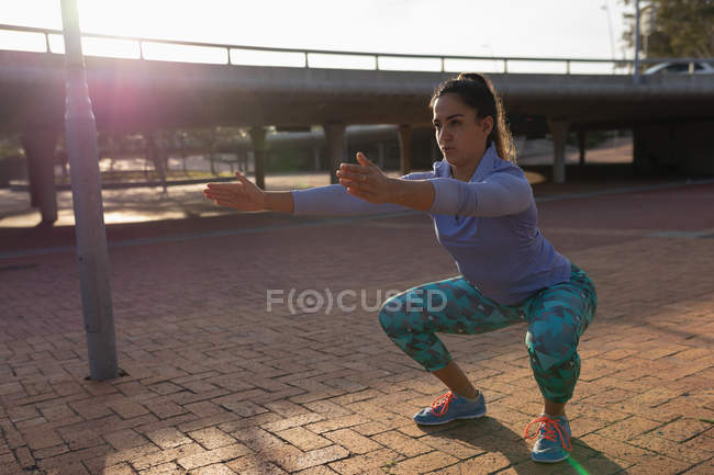 Vista frontal close-up de uma jovem caucasiana vestindo roupas esportivas agachamento com os braços estendidos na frente dela durante um treino em um parque — Fotografia de Stock