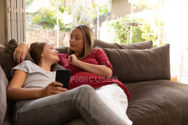 Vorderansicht einer jungen kaukasischen Schwangeren, die mit ihrer Zwillingstochter spricht, die auf einem Sofa im Wohnzimmer sitzt und ein Smartphone in der Hand hält — Stockfoto