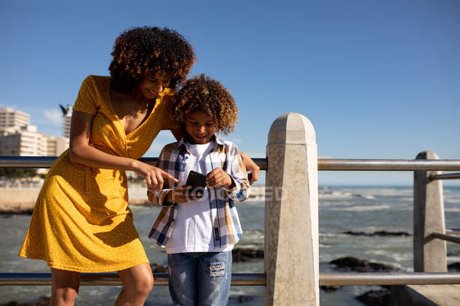 Vista frontal de uma jovem mulher de raça mista e seu filho pré-adolescente desfrutando de tempo juntos junto ao mar, de pé e usando um smartphone em um dia ensolarado — Fotografia de Stock