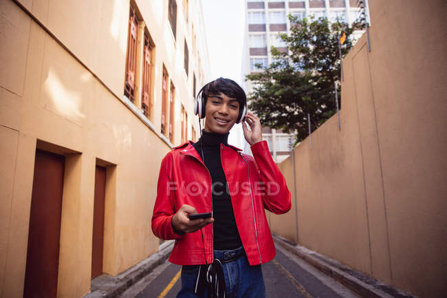 Retrato de un joven transexual de raza mixta de moda en la calle, usando un teléfono inteligente con auriculares sonriendo a la cámara - foto de stock