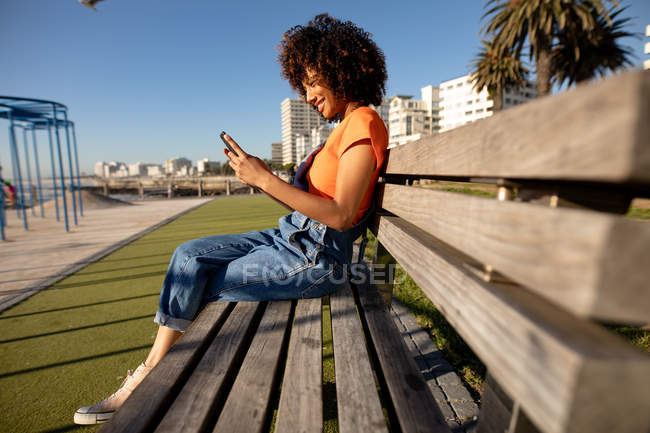 Vista lateral de una joven mestiza sentada en un banco junto a un parque infantil, usando un smartphone en un día soleado - foto de stock