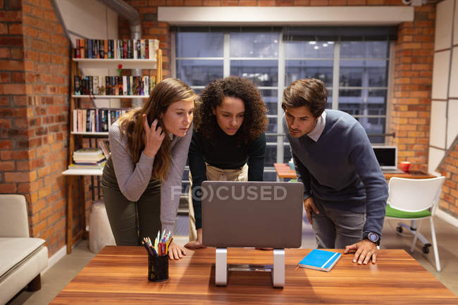 Фронт-вью крупным планом молодой смешанной расы женщина и молодая кавказская женщина и мужчина, стоящие в дискуссии вокруг ноутбука компьютер, работающий вместе в офисе творческого бизнеса — стоковое фото