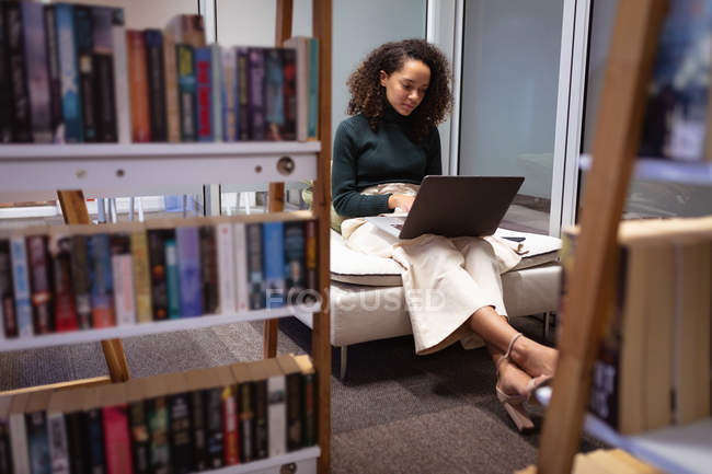 Vista frontal de uma jovem mulher de raça mista sentada em uma sala de estar trabalhando no computador portátil no escritório de um negócio criativo — Fotografia de Stock
