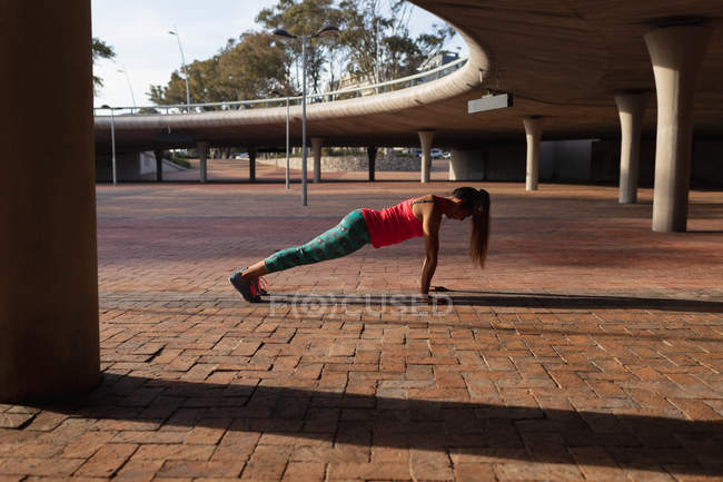 Vista lateral de una joven mujer caucásica con ropa deportiva haciendo flexiones durante un entrenamiento en un día soleado en un parque - foto de stock