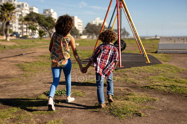 Vista trasera de una joven mestiza y su hijo preadolescente disfrutando del tiempo juntos caminando a un parque infantil junto al mar y tomados de la mano en un día soleado, con columpios en el fondo - foto de stock