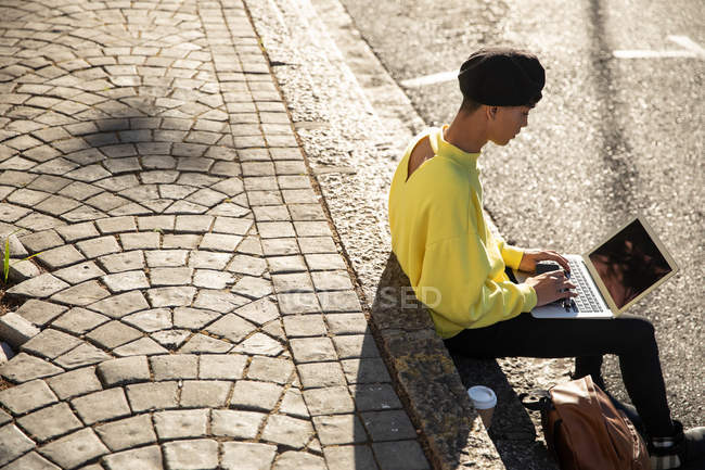 Вид збоку модного молодого змішаної раси транссексуал дорослий на вулиці, використовуючи ноутбук комп'ютер сидить на кроках — стокове фото