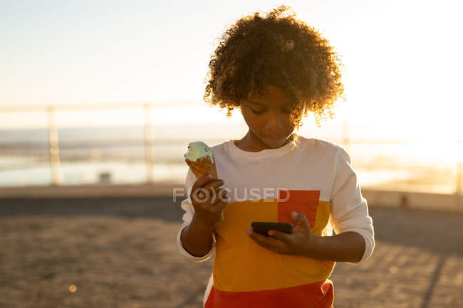 Front view of a pre-teen boy holding an ice cream and looking down at a smartphone by the sea, backlit by the setting sun — Stock Photo