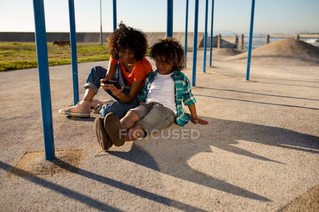 Вид спереди на молодую женщину смешанной расы и ее сына-подростка, наслаждающуюся временем вместе, играющую на детской площадке у моря, сидящую на земле у скалолазания и делающую селфи в солнечный день — стоковое фото