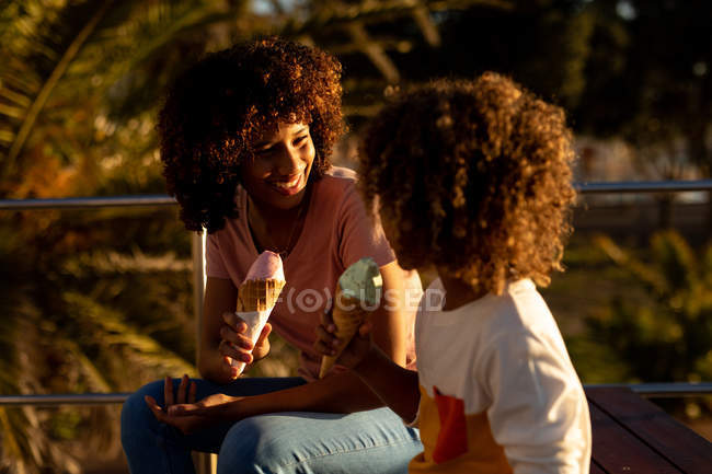 Vista frontal de una mujer de raza mixta y su hijo preadolescente disfrutando del tiempo juntos junto al mar, sonriéndose, sentándose y comiendo helado en un día soleado - foto de stock