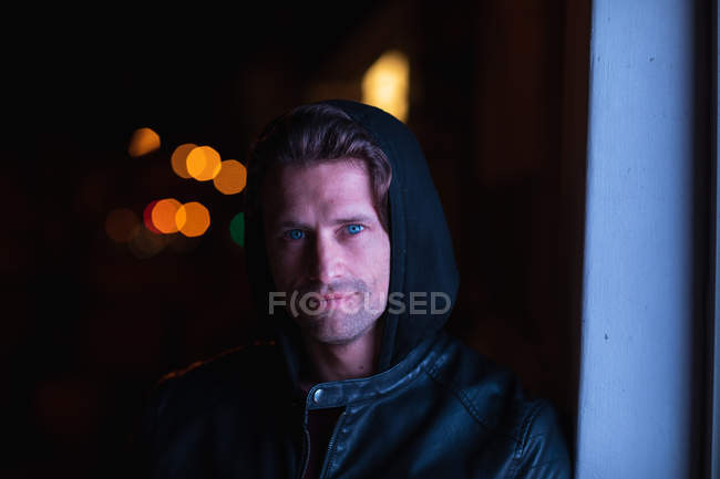 Портрет усміхнений молодий Кавказький чоловік одягнений в балахон дивлячись на камеру спираючись на стіну на міській вулиці вночі — стокове фото