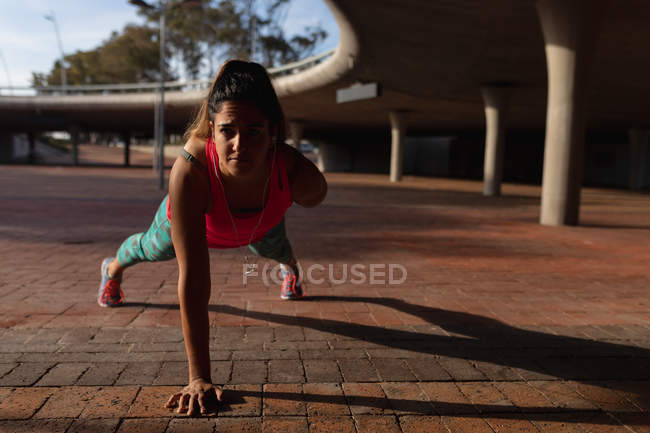 Vista frontal close-up de uma jovem caucasiana vestindo roupas esportivas fazendo flexões em um braço durante um treino em um dia ensolarado em um parque — Fotografia de Stock