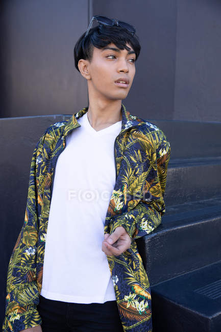 Vista frontal de um jovem adulto transexual de raça mista na moda na rua, em pé junto a uma escada e uma parede cinza — Fotografia de Stock